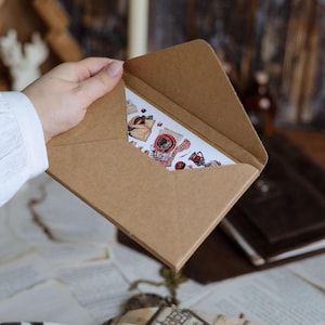 K&ouml;nnte beinhalten: Ein brauner Kraftpapierumschlag enth&auml;lt eine Karte mit verschiedenen Aufklebern. Die Karte zeigt Bilder von Kaffee, B&uuml;chern und Elementen zum Thema Vintage. Die Szene spielt auf einem Tisch mit verstreuten Papieren und einem unscharfen Hintergrund.