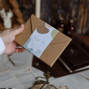 K&ouml;nnte beinhalten: Ein braunes Papierpaket mit einem wei&szlig;en Etikett mit der Aufschrift "Thank You for your order". Das Etikett hat abstrakte Formen in Braun, Gr&uuml;n und Grau. Das Paket wird von einer Person gehalten.