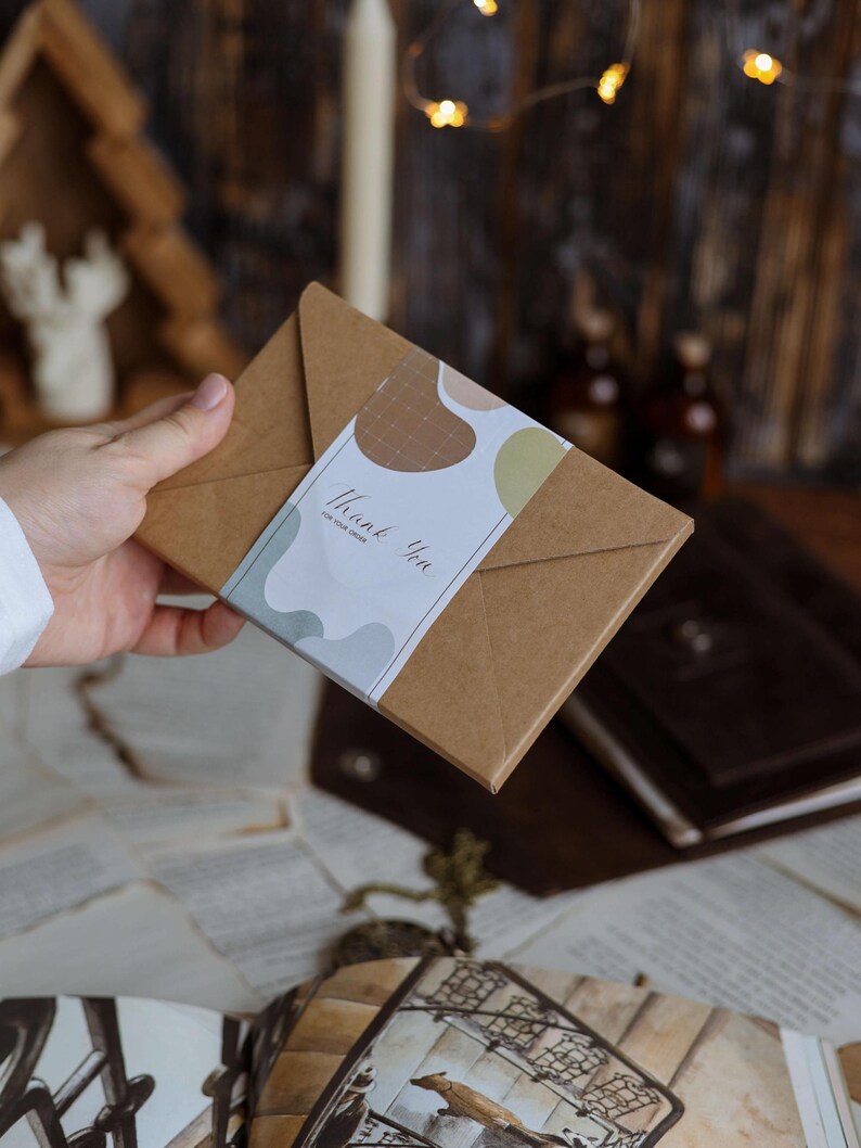 K&ouml;nnte beinhalten: Ein braunes Papierpaket mit einem wei&szlig;en Etikett mit der Aufschrift "Thank You for your order". Das Etikett hat abstrakte Formen in Braun, Gr&uuml;n und Grau. Das Paket wird von einer Person gehalten.