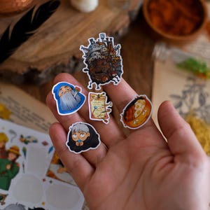 May include: A collection of colorful stickers featuring illustrations of characters and scenes from an animated film. The stickers include images of a castle, a wizard, a map, a character wrapped in a blanket, and a plate of food.