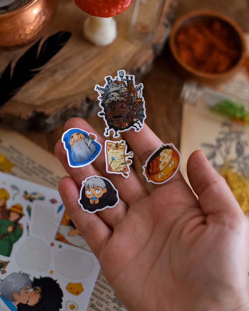 May include: A collection of colorful stickers featuring illustrations of characters and scenes from an animated film. The stickers include images of a castle, a wizard, a map, a character wrapped in a blanket, and a plate of food.