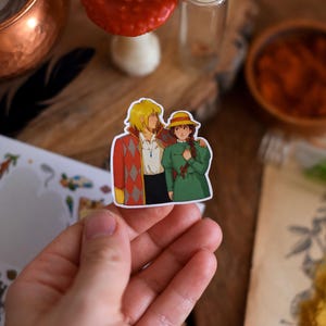 May include: A colorful sticker featuring two anime characters. One character has blonde hair and is wearing a red and orange diamond-patterned jacket. The other character has brown hair and is wearing a green dress and a yellow hat. The sticker is held in a hand.