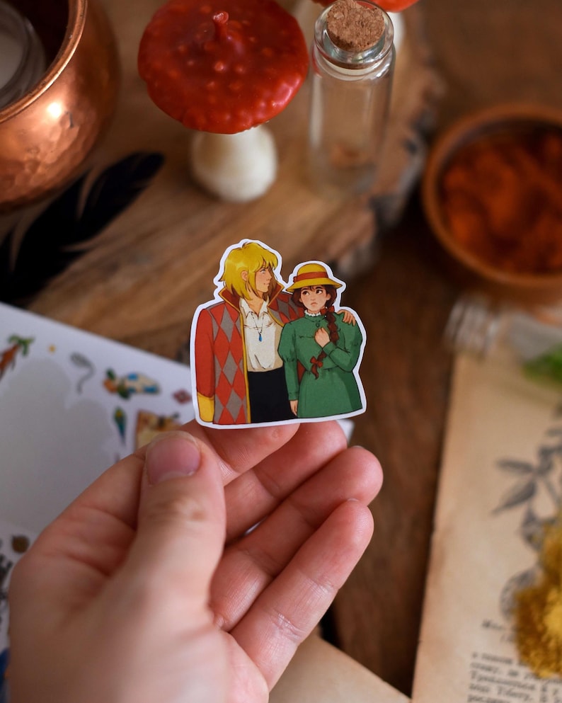May include: A colorful sticker featuring two anime characters. One character has blonde hair and is wearing a red and orange diamond-patterned jacket. The other character has brown hair and is wearing a green dress and a yellow hat. The sticker is held in a hand.