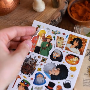 May include: A sheet of colorful stickers featuring characters and scenes from an animated film. The stickers depict various characters, a castle, and food items. The stickers are held by a hand against a wooden surface.