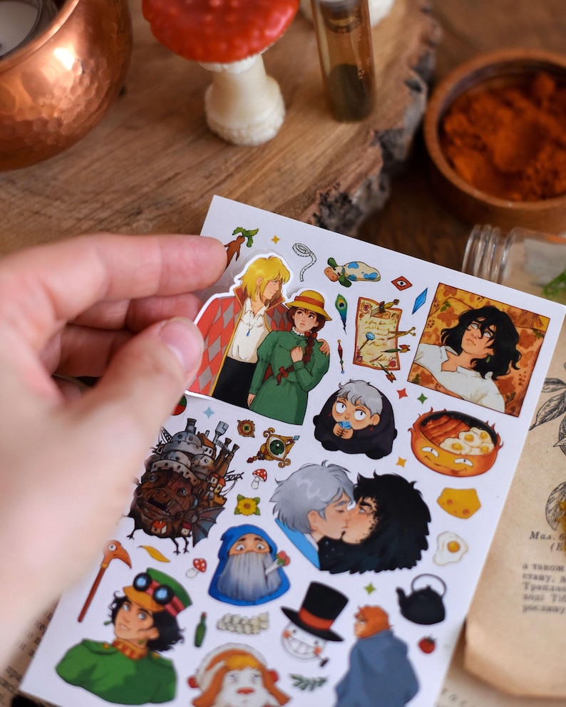 May include: A sheet of colorful stickers featuring characters and scenes from an animated film. The stickers depict various characters, a castle, and food items. The stickers are held by a hand against a wooden surface.