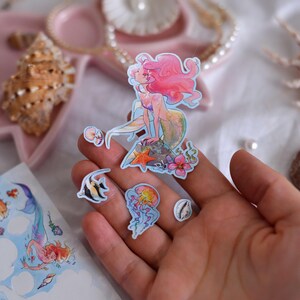 Puede incluir: Un conjunto de pegatinas con tem&aacute;tica de sirenas. Las pegatinas presentan una sirena con pelo rosa, un pez, una medusa y otros dise&ntilde;os marinos. Las pegatinas son coloridas y tienen un estilo de acuarela.