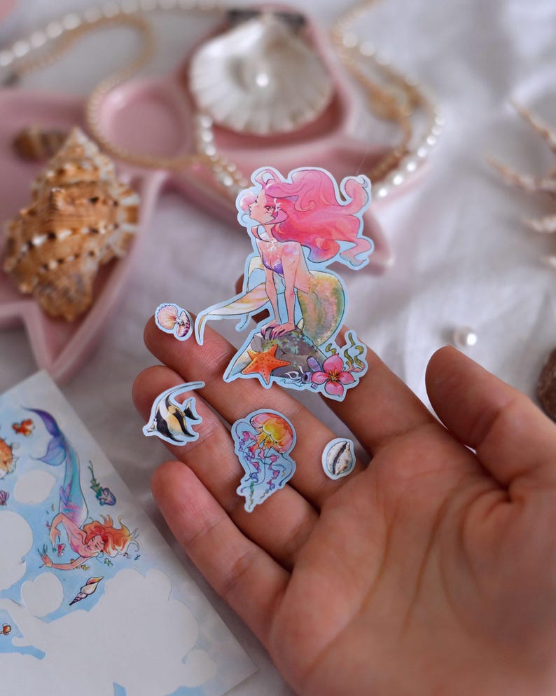 Puede incluir: Un conjunto de pegatinas con tem&aacute;tica de sirenas. Las pegatinas presentan una sirena con pelo rosa, un pez, una medusa y otros dise&ntilde;os marinos. Las pegatinas son coloridas y tienen un estilo de acuarela.