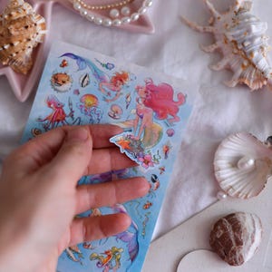 Puede incluir: Una hoja de pegatinas con tem&aacute;tica de sirenas, con ilustraciones coloridas de sirenas, criaturas marinas y plantas acu&aacute;ticas. Las pegatinas se sostienen en una mano, con conchas marinas y un collar de perlas en el fondo.