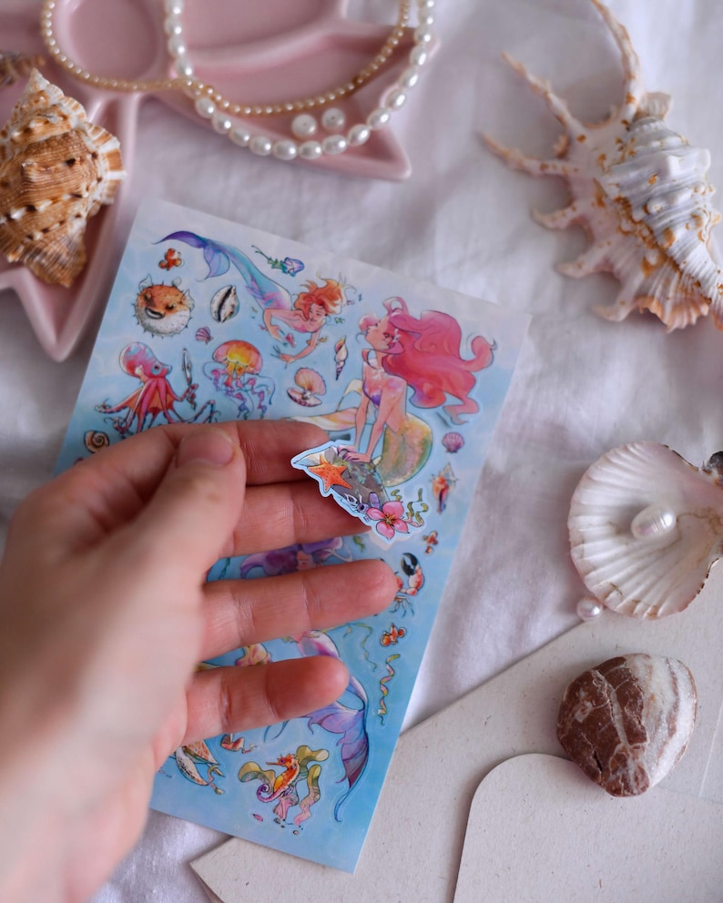 Puede incluir: Una hoja de pegatinas con tem&aacute;tica de sirenas, con ilustraciones coloridas de sirenas, criaturas marinas y plantas acu&aacute;ticas. Las pegatinas se sostienen en una mano, con conchas marinas y un collar de perlas en el fondo.