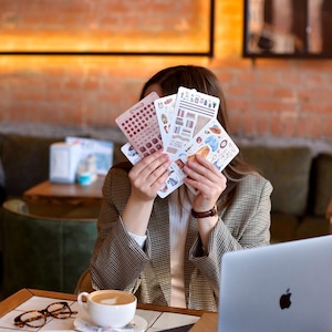 Puede incluir: Una persona sostiene una colecci&oacute;n de hojas de pegatinas con varios dise&ntilde;os, incluyendo calendarios e ilustraciones. La persona lleva una blazer a cuadros marr&oacute;n y beige. Una computadora port&aacute;til, caf&eacute; y postre est&aacute;n sobre la mesa.