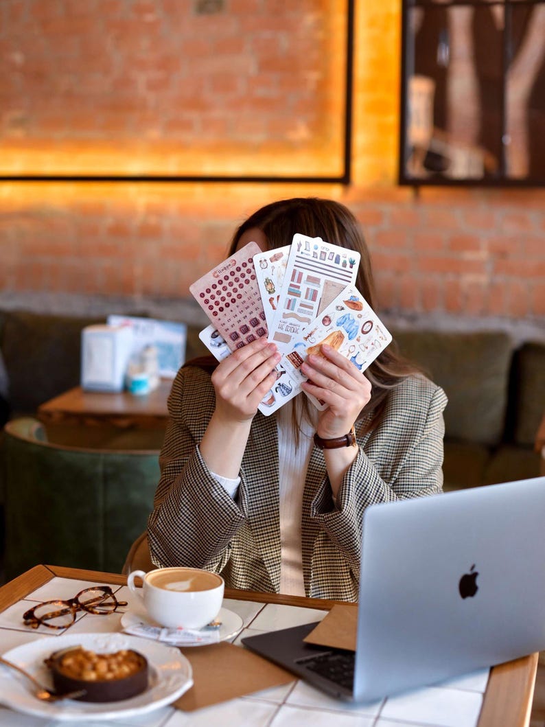 Puede incluir: Una persona sostiene una colecci&oacute;n de hojas de pegatinas con varios dise&ntilde;os, incluyendo calendarios e ilustraciones. La persona lleva una blazer a cuadros marr&oacute;n y beige. Una computadora port&aacute;til, caf&eacute; y postre est&aacute;n sobre la mesa.