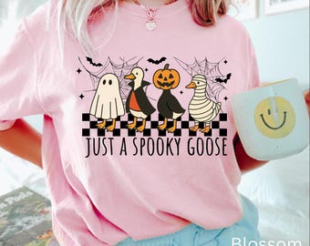 May include: Pink t-shirt with a Halloween-themed graphic. The design features four cartoon geese dressed as a ghost, vampire, jack-o'-lantern, and mummy, with the text "JUST A SPOOKY GOOSE". Bats and spiderwebs complete the design.