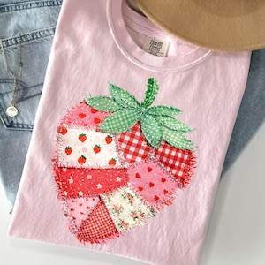 Peut inclure: T-shirt rose clair avec un motif de fraise en patchwork. La fraise est composée de divers tissus à motifs rouges et roses, avec une feuille verte à carreaux et à pois. Le mot "Blossom" est en bas.