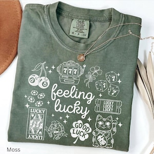 Può includere: T-shirt verde muschio con la scritta "feeling lucky" e vari simboli fortunati bianchi, tra cui dadi, carte da gioco e un gatto portafortuna. La maglietta ha un girocollo e maniche corte.