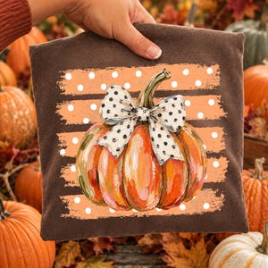 May include: A dark brown fabric square featuring a central graphic of an orange pumpkin with visible brushstroke textures. The pumpkin is adorned with a white bow featuring black polka dots, set against a background of horizontal orange stripes with white dots.