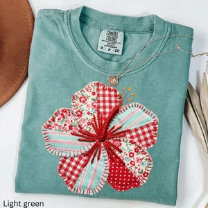Könnte beinhalten: Hellgrünes T-Shirt mit einem großen, applizierten Blumendesign. Die Blume hat rote Vichy-, Blumen- und Tupfen-Stoffflicken mit einem rot gestickten Zentrum. Eine silberne Halskette mit Anhänger liegt auf dem Shirt.