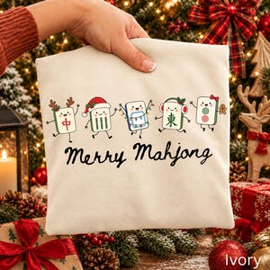 May include: An ivory-colored sweatshirt featuring a graphic of five cartoon Mahjong tiles decorated for the holidays, including reindeer antlers, a Santa hat, string lights, earmuffs, and a bow. The text Merry Mahjong is printed in black below.