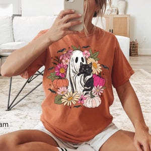 May include: A burnt orange short-sleeve t-shirt featuring a graphic print of a white ghost holding a black cat, surrounded by colorful flowers, pumpkins, and small black bats. The design is centered on the chest of the garment.
