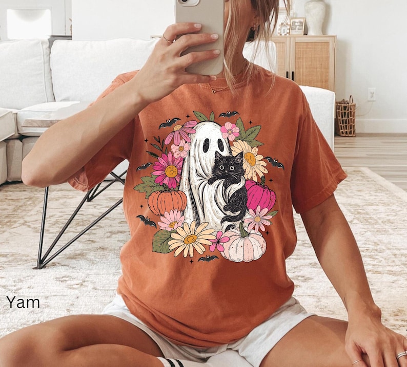 May include: A burnt orange short-sleeve t-shirt featuring a graphic print of a white ghost holding a black cat, surrounded by colorful flowers, pumpkins, and small black bats. The design is centered on the chest of the garment.