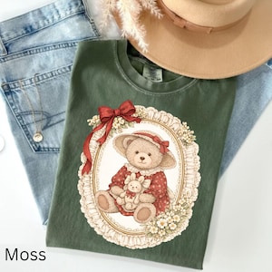 May include: A moss green t-shirt featuring a framed illustration of a teddy bear wearing a hat and holding a stuffed bunny. The bear is dressed in a red dress with white lace trim. A red bow adorns the top of the frame. A tan hat and blue jeans are also in the image.