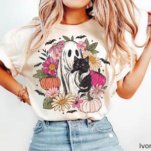 May include: An ivory t-shirt featuring a graphic print of a white ghost holding a black cat, surrounded by colorful flowers, green leaves, orange and pink pumpkins, and small black bats. The design is centered on the front of the garment.