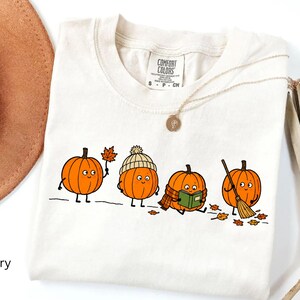 May include: An ivory t-shirt featuring a graphic of four orange pumpkins with faces. One pumpkin holds a leaf, one wears a beanie, one reads a green book while wearing a scarf, and one holds a broom. The shirt has a Comfort Colors brand label.