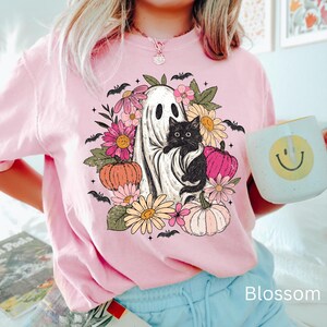 May include: A light pink t-shirt featuring a graphic print of a white ghost holding a black kitten, surrounded by colorful flowers, pumpkins, and small black bats. The design includes pink, orange, and yellow floral elements and pumpkins.