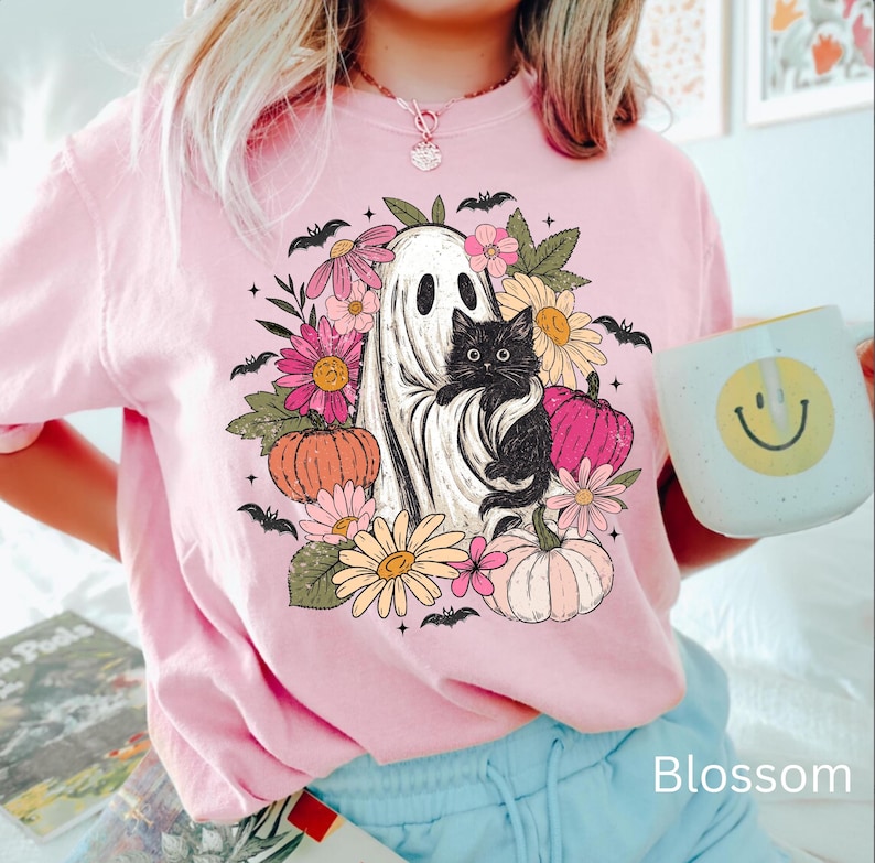 May include: A light pink t-shirt featuring a graphic print of a white ghost holding a black kitten, surrounded by colorful flowers, pumpkins, and small black bats. The design includes pink, orange, and yellow floral elements and pumpkins.