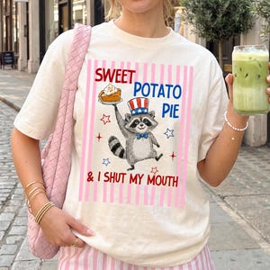 以下が含まれることがあります： アイボリー色のTシャツ。愛国的な帽子をかぶったアライグマがサツマイモパイを持っているグラフィック。テキストは「Sweet Potato Pie & I Shut My Mouth」と赤と青で書かれています。