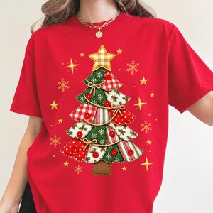 May include: A red t-shirt featuring a graphic of a Christmas tree made of various fabric patterns including plaid, polka dots, and snowflakes. The tree is decorated with red buttons, small bows, and a yellow gingham star on top, surrounded by gold stars.