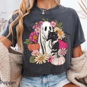 May include: A charcoal gray short-sleeve t-shirt featuring a graphic print of a white ghost holding a black cat, surrounded by colorful flowers, green leaves, small bats, and orange and pink pumpkins arranged in a circular pattern.