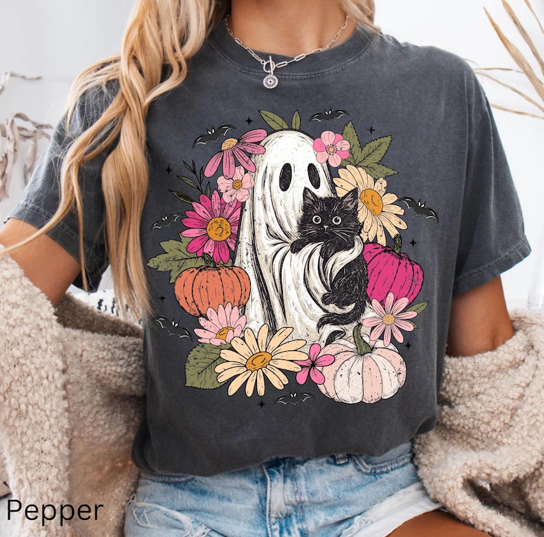 May include: A charcoal gray short-sleeve t-shirt featuring a graphic print of a white ghost holding a black cat, surrounded by colorful flowers, green leaves, small bats, and orange and pink pumpkins arranged in a circular pattern.