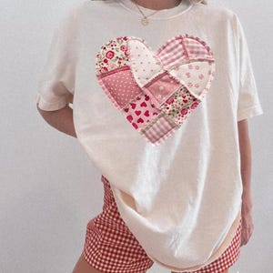 May include: Ivory-coloured t-shirt with a patchwork heart design in pink and red hues. The heart is made from various floral and patterned fabrics. The model is wearing red and white checked shorts. The word "Ivory" is printed on the bottom right.