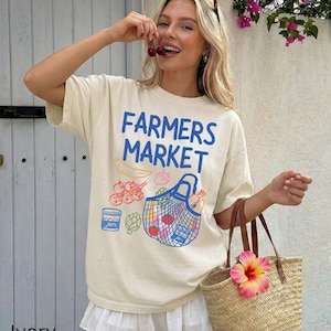 Könnte beinhalten: Elfenbeinfarbenes T-Shirt mit dem blauen Text "FARMERS MARKET". Das Shirt hat eine Grafik einer Einkaufstasche mit Produkten. Eine Stroh-Tragetasche mit einer rosa Blume wird in der Hand gehalten.