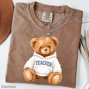 May include: A brown short-sleeve t-shirt featuring a graphic of a brown teddy bear wearing a white hooded sweatshirt with the word TEACHER printed in blue across the chest. The shirt has a crew neckline and a Comfort Colors brand label inside.