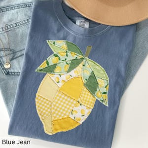 May include: A blue jean t-shirt with a patchwork lemon design. The lemon is made of yellow and green fabric with lemon and gingham patterns. A tan hat and blue jeans are in the background.