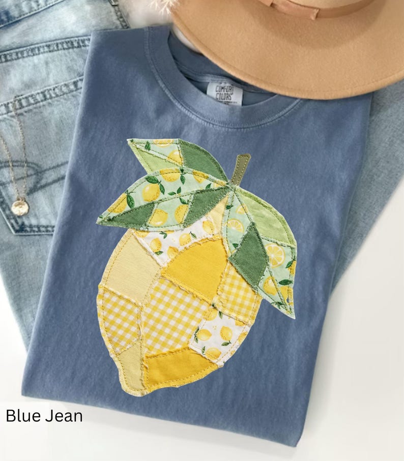 May include: A blue jean t-shirt with a patchwork lemon design. The lemon is made of yellow and green fabric with lemon and gingham patterns. A tan hat and blue jeans are in the background.