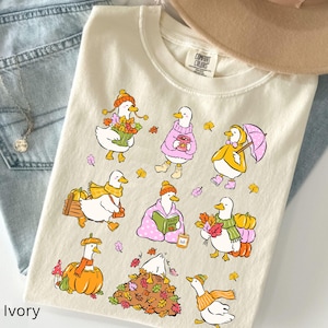 Puede incluir: Una camiseta de color marfil con un estampado gráfico de nueve patos blancos con diversos atuendos otoñales, que incluyen sostener calabazas, usar gorros y bufandas de punto, leer un libro y jugar en un montón de hojas de otoño coloridas.