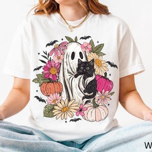 May include: A white t-shirt featuring a graphic print of a ghost holding a black cat, surrounded by colorful flowers, pumpkins, and small black bats. The design includes pink, orange, and yellow floral elements with green leaves and pumpkins.