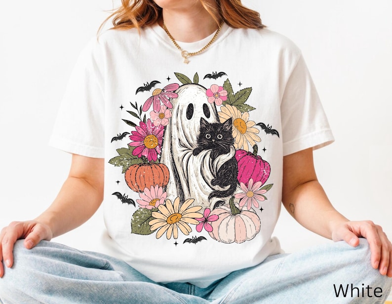 May include: A white t-shirt featuring a graphic print of a ghost holding a black cat, surrounded by colorful flowers, pumpkins, and small black bats. The design includes pink, orange, and yellow floral elements with green leaves and pumpkins.