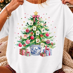 May include: A white t-shirt featuring a graphic of a green Christmas tree decorated with Mahjong tiles, red ornaments, and a gold star. The tree sits in a blue and white patterned ceramic pot, surrounded by three wrapped gift boxes at the base.