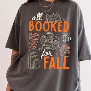 以下が含まれることがあります： チャコールグレーの半袖Tシャツ。オレンジとクリーム色の「all BOOKED for FALL」という文字がプリントされています。グラフィックには、積み重ねられた本、開いた本、コーヒーカップ、カボチャ、キャンドル、小さな秋の葉のイラストが描かれています。