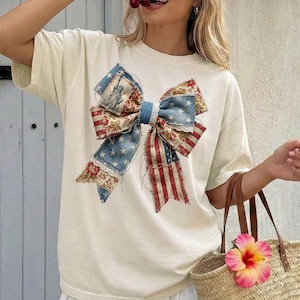 Puede incluir: Camiseta de color marfil con un gran diseño de lazo con patrones patrióticos. El lazo incorpora elementos de la bandera estadounidense, estampados florales y la Estatua de la Libertad. La camiseta tiene un corte holgado y mangas cortas.