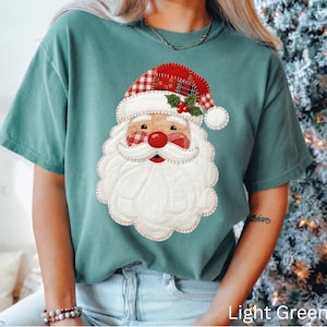 Op de afbeelding: Een lichtgroen t-shirt met korte mouwen en een grote, centrale afbeelding van een kerstmangezicht. Het ontwerp heeft een muts met een rood-wit ruitpatroon, een witte doorgestikte baard en een rode neus, alles afgewerkt met decoratieve stiksels.