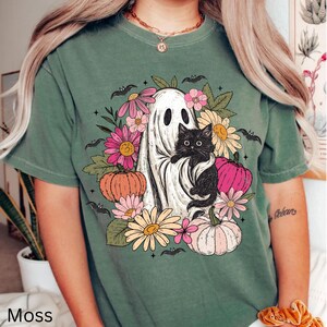 May include: A moss green short-sleeve t-shirt featuring a graphic print of a white ghost holding a black cat, surrounded by colorful flowers, pumpkins, and small bats. The design includes pink, orange, and yellow floral elements and pumpkins.
