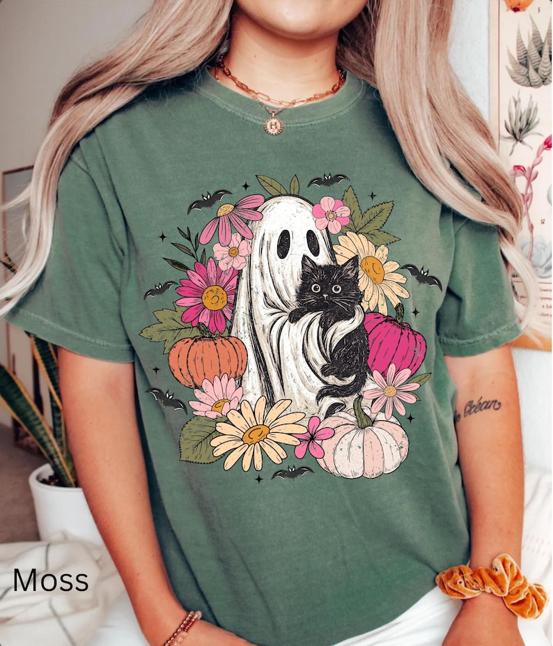 May include: A moss green short-sleeve t-shirt featuring a graphic print of a white ghost holding a black cat, surrounded by colorful flowers, pumpkins, and small bats. The design includes pink, orange, and yellow floral elements and pumpkins.