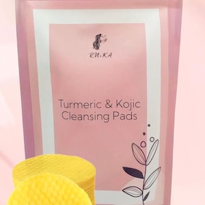 May include: A pink resealable pouch with a white label that says "R.U.K.A Turmeric & Kojic Cleansing Pads". The label also features a stylized floral design.  A stack of yellow cleansing pads are in the foreground.