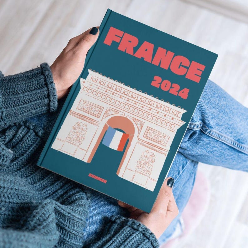 Może przedstawiać: Niebiesko-zielony notes z białą ilustracją Łuku Triumfalnego w Paryżu, Francja. Notes ma napis "FRANCE 2024" w czerwono-białych literach.