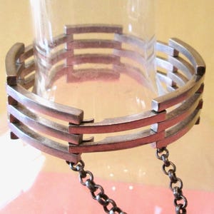 May include: A silver-toned bracelet with a geometric design. The bracelet features four curved, parallel bars connected by small rectangular links. A delicate chain hangs from the bottom. The bracelet is displayed on a clear glass object.