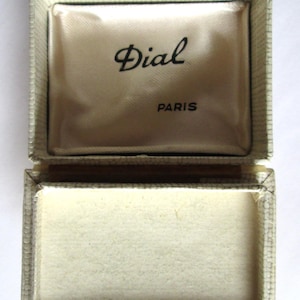May include: Open vintage jewelry box with a cream-colored interior and exterior. The box is rectangular with a hinged lid. The inside lid has the word "Dial" in cursive above "PARIS". The box is empty.